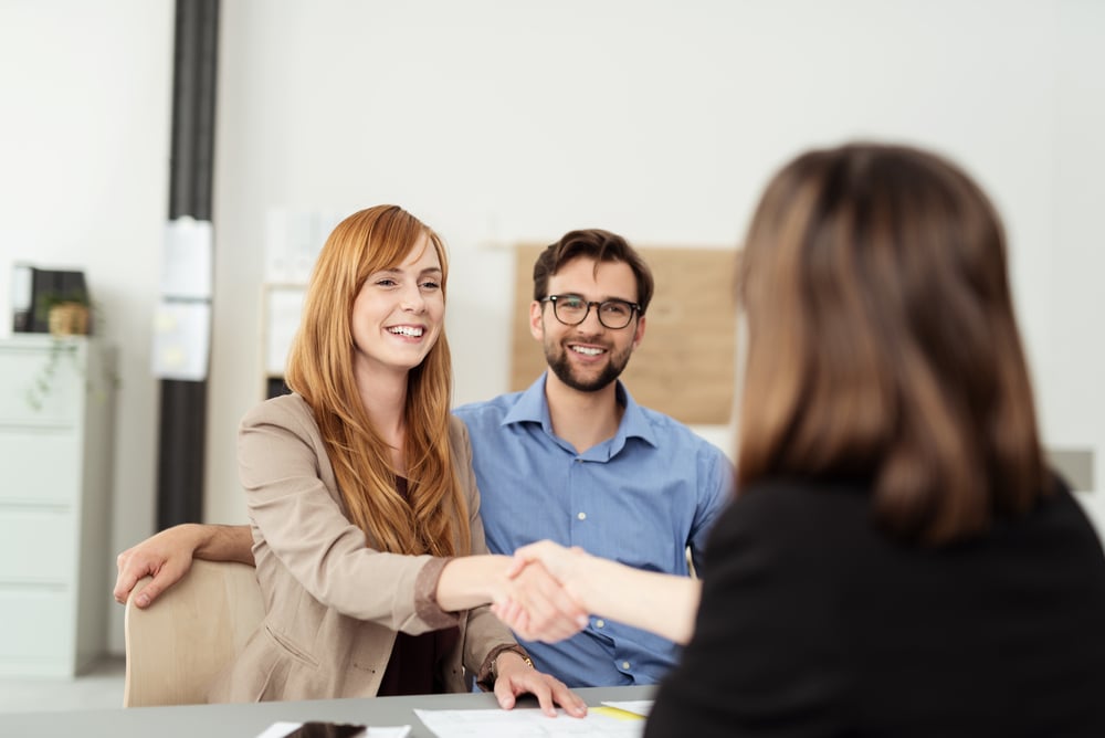 Happy young couple meeting with a broker in her office leaning over the desk to shake hands, view from behind the female agent Happy young couple meeting with a broker in her office leaning over the desk to shake hands, view from behind the female agent